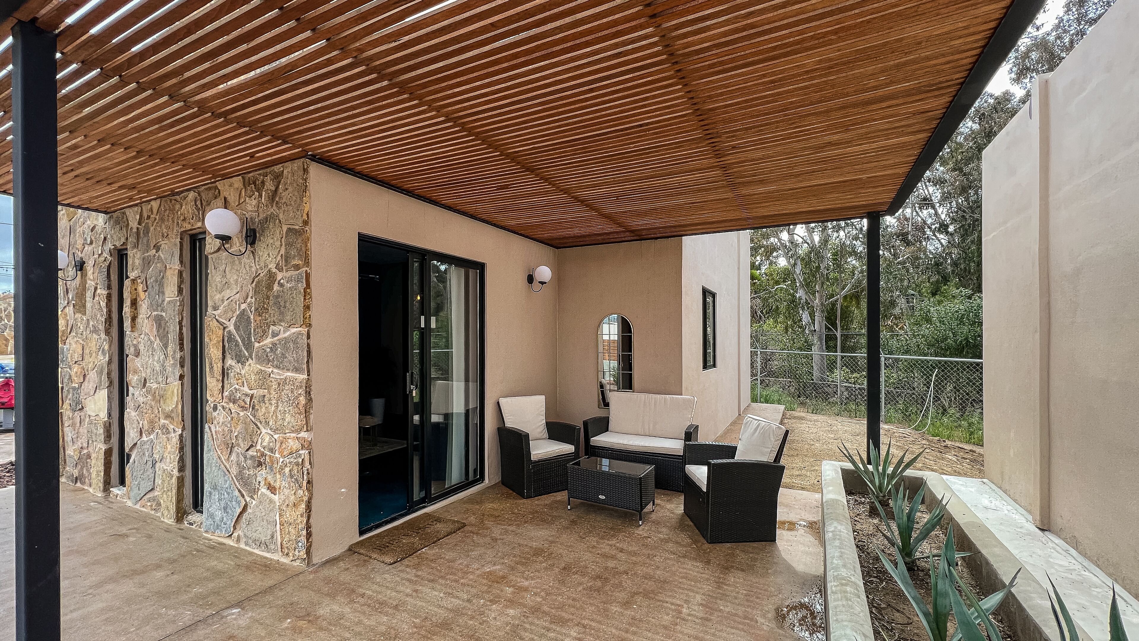 Ein überdachter Patio-Bereich mit Steinwänden, einer Glastür zum Innenbereich und Gartenmöbeln.