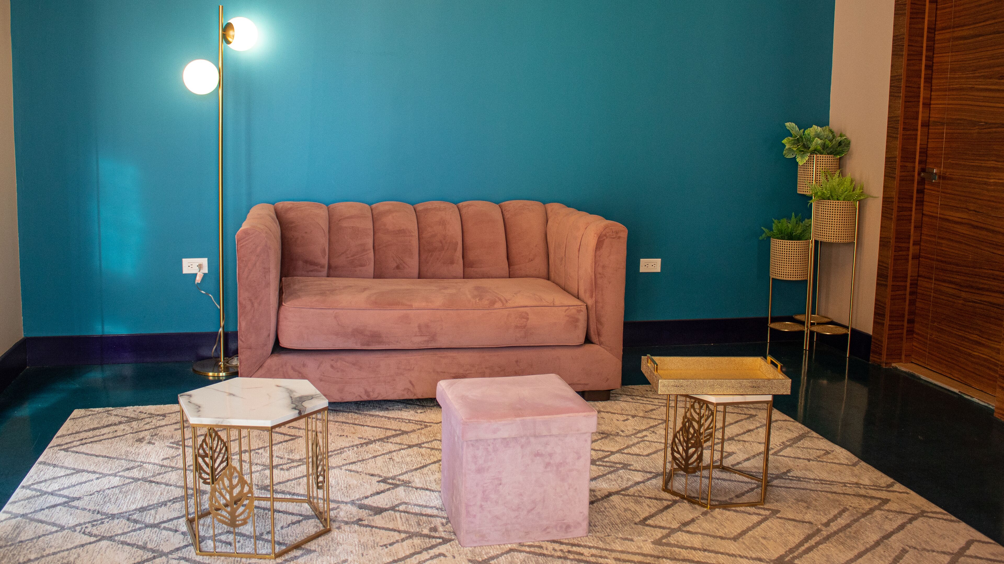A room with a teal wall, a pink sofa, a marble coffee table, and two gold-framed side tables.