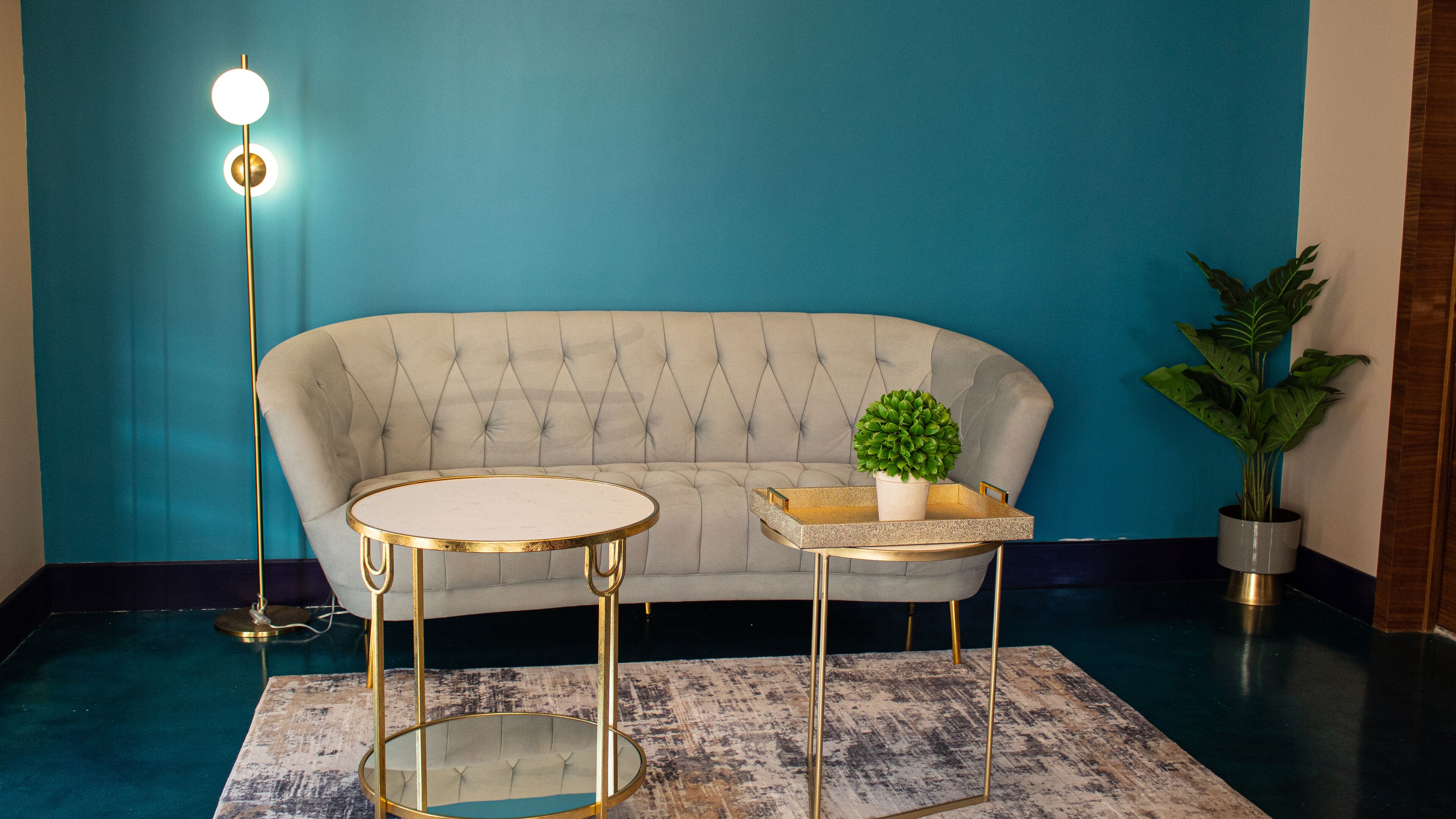 Une pièce comprenant un canapé capitonné, deux tables encadrées d’or, un lampadaire et une plante en pot.