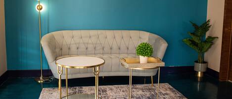 A room with a tufted sofa, two gold-framed tables, a floor lamp, and a potted plant.
