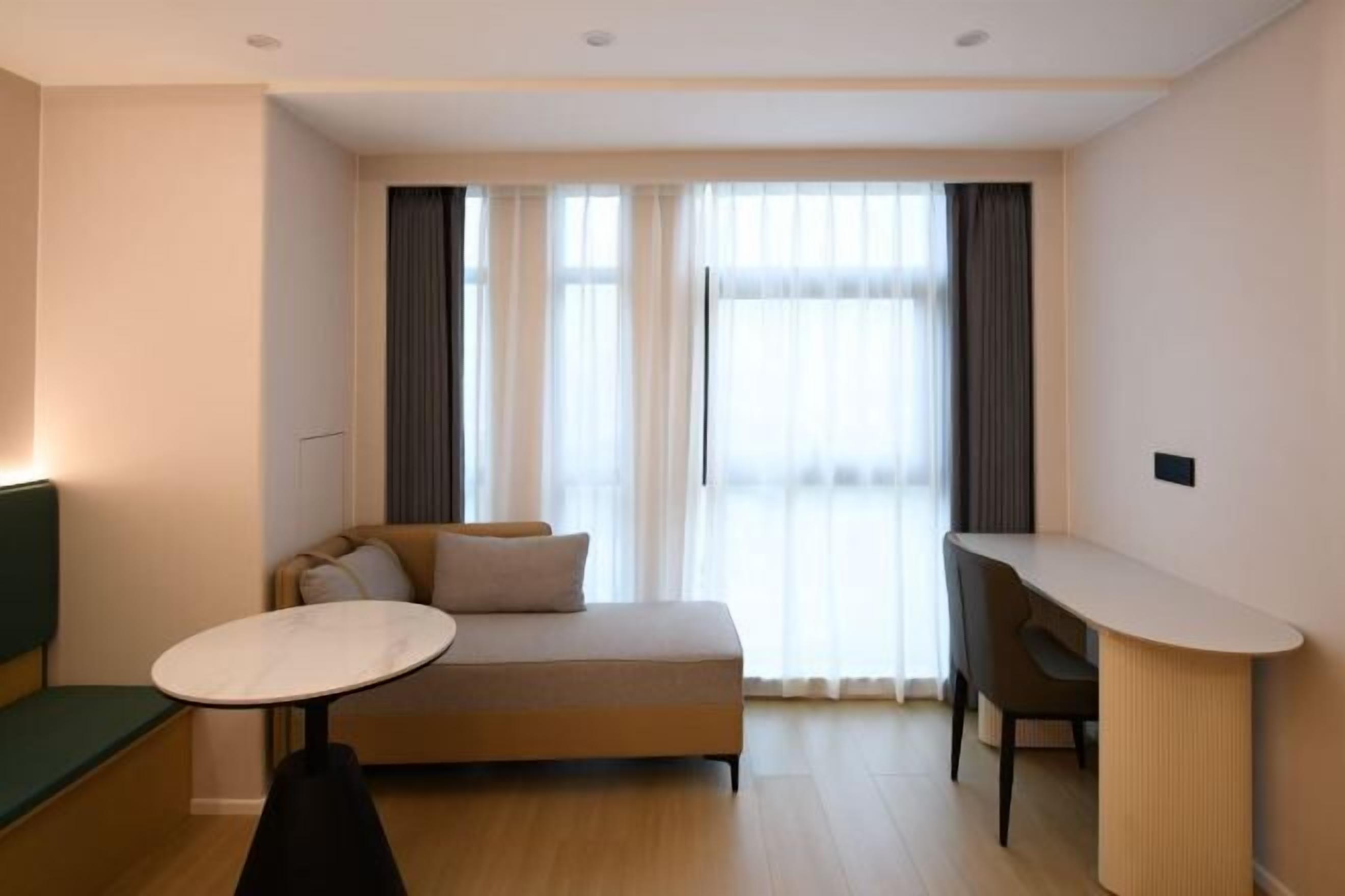 Una habitación de hotel moderna con sofá, una pequeña mesa redonda, escritorio y ventanas grandes con cortinas.