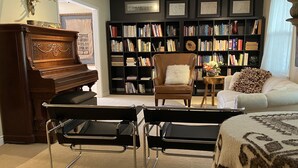 Een woonkamer met een vleugelpiano, een boekenplank, een bruine leren fauteuil, een witte bank en een kamerbreed tapijt met dessin.
