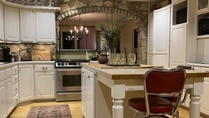 Een keuken met witte kastjes, een stenen boog, een centraal eiland met krukken, een kroonluchter en een kamerbreed tapijt met een patroon.