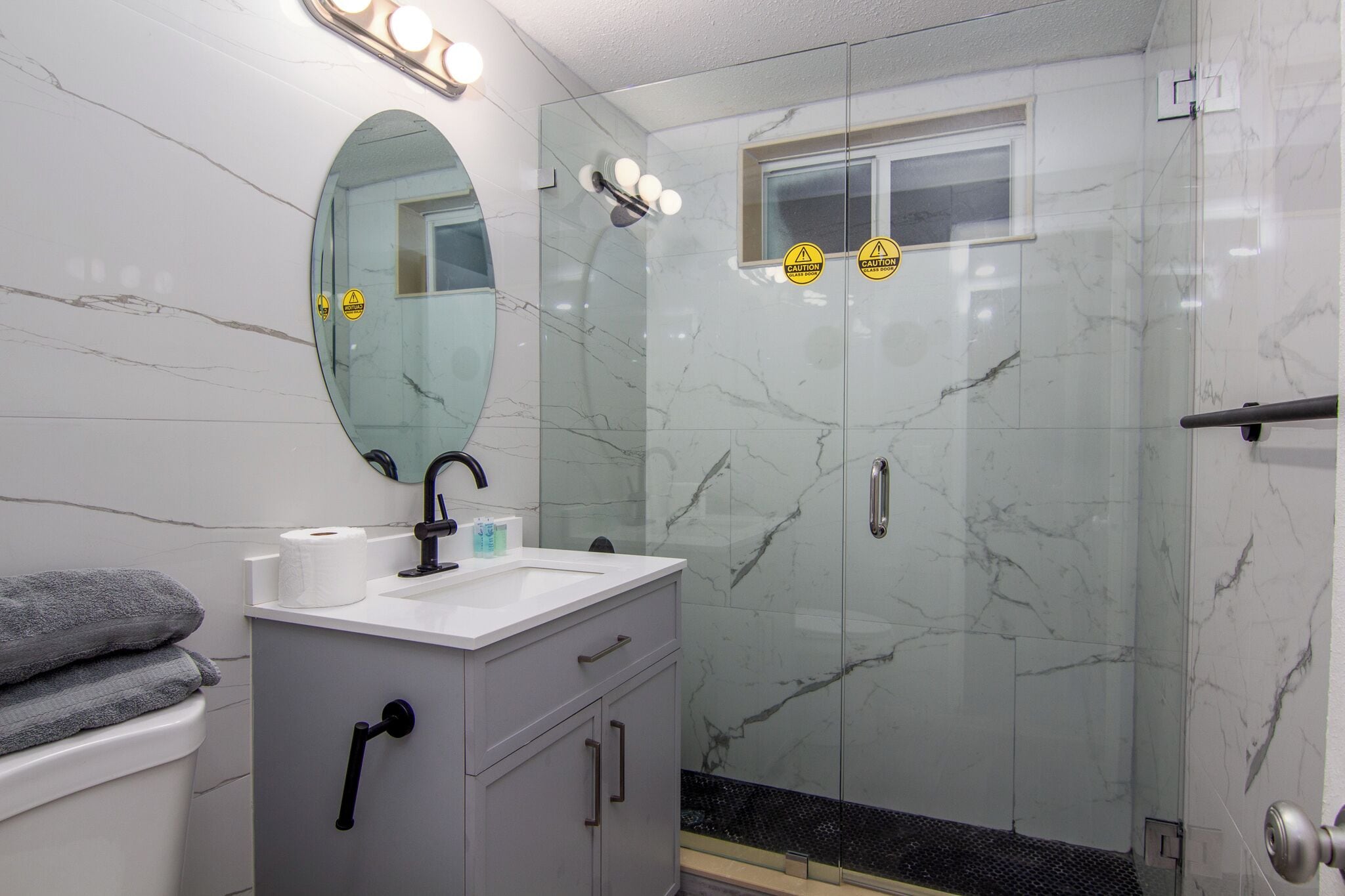 A bathroom with a marble wall, a glass shower enclosure, a sink with a mirror, and a toilet.