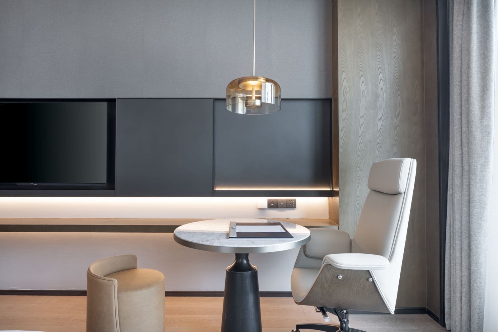 A modern living room with a flat-screen TV, a round table with a book, a chair, and a pendant light.