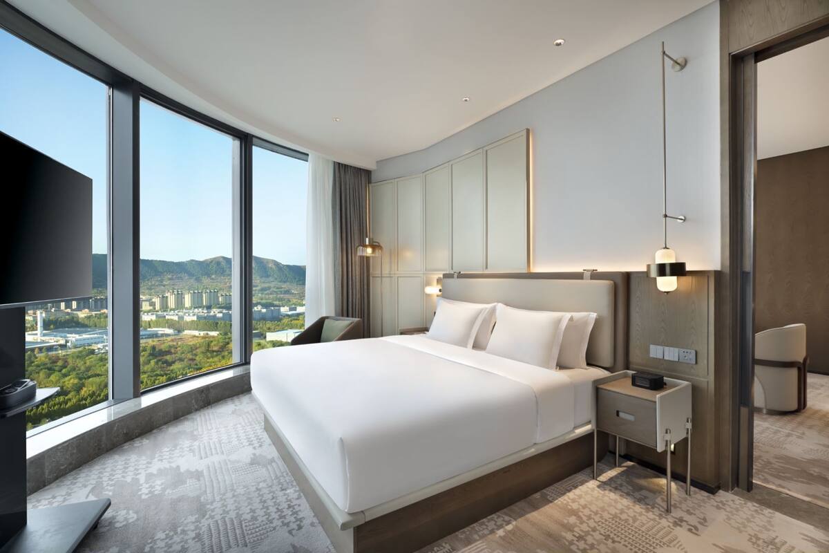 Een moderne hotelkamer met een groot bed, een flatscreen-tv, een nachtkastje en uitzicht op een stadsbeeld door grote ramen.