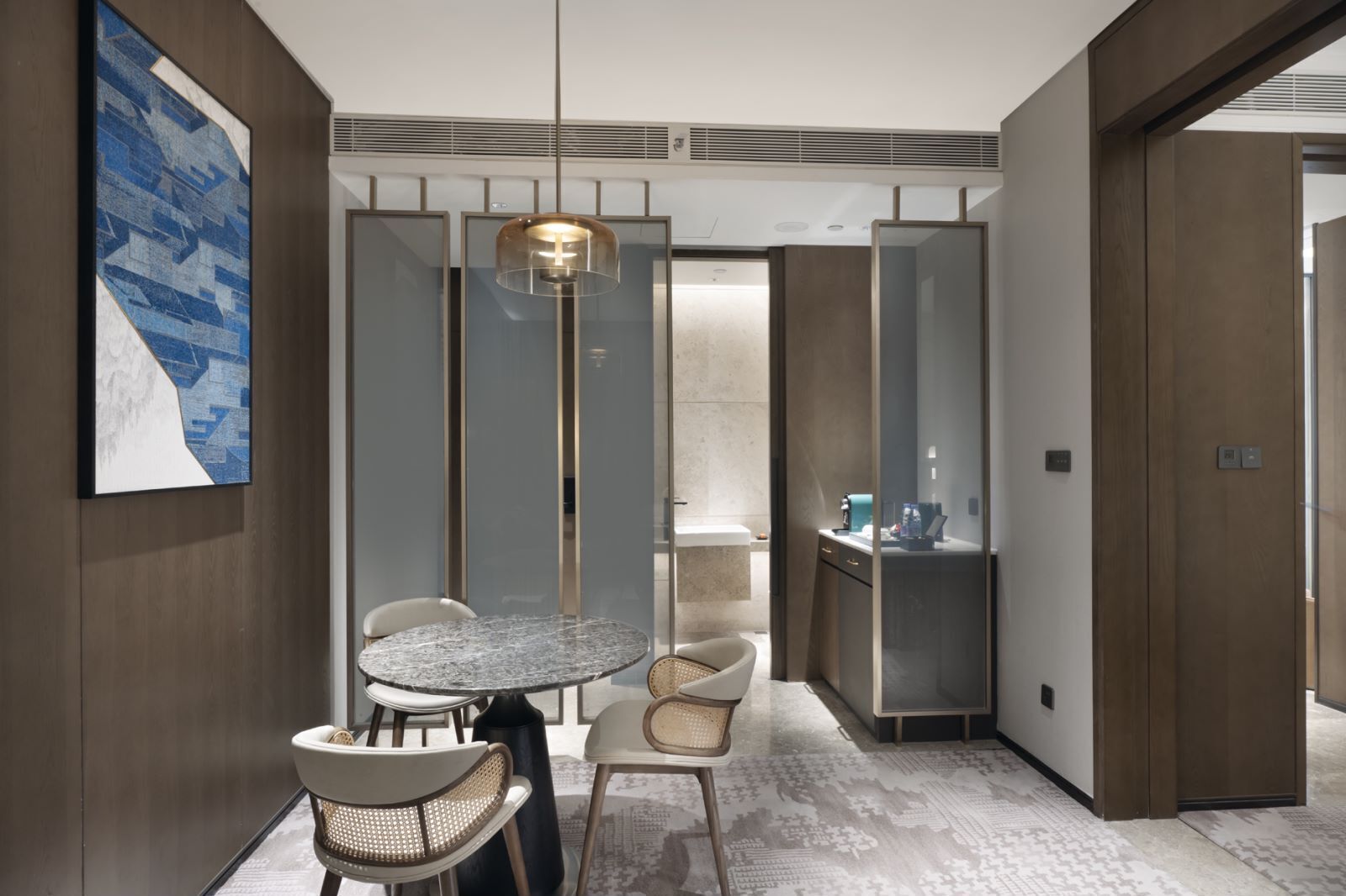 Een moderne hotelkamer met een ronde marmeren tafel, twee stoelen, een hanglamp, een groot abstract schilderij, een glazen scheidingswand, een minibar en een houten deur.