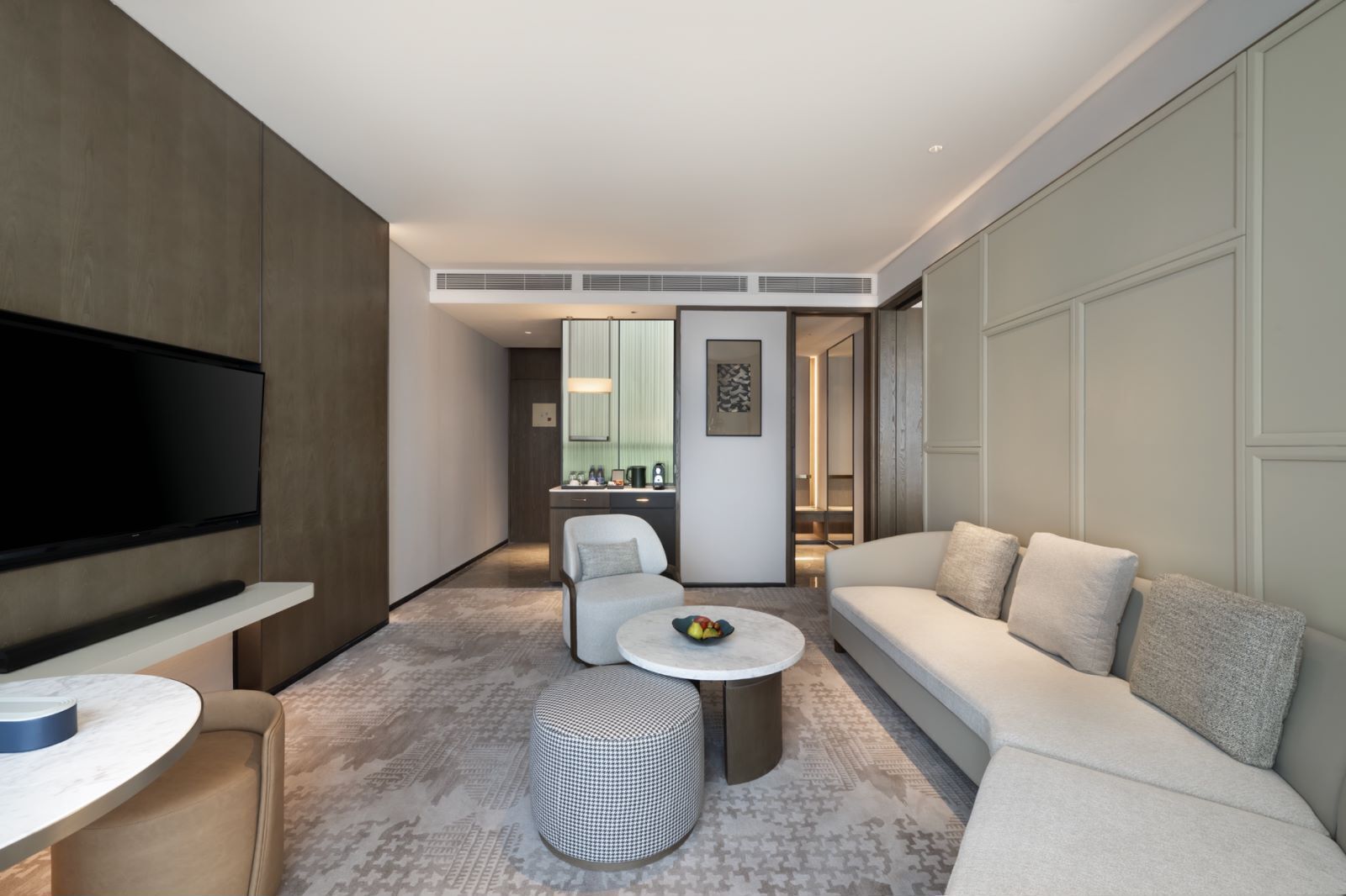 A modern hotel room with a flat-screen TV, a sofa, a dining table, and a small round table with a bowl of fruit.
