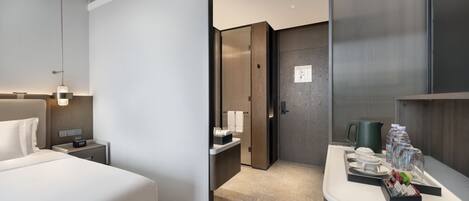Una habitación de hotel moderna con una cama grande, una mesita de noche y un escritorio pequeño con una lámpara. Se ve un baño a través de una puerta abierta.