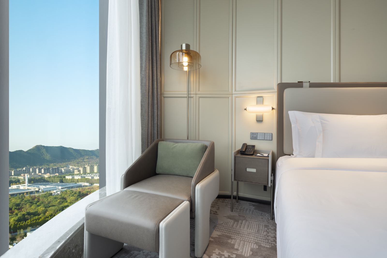 Habitación de hotel con cama, una silla, una mesita de noche con lámpara y vistas a la ciudad.