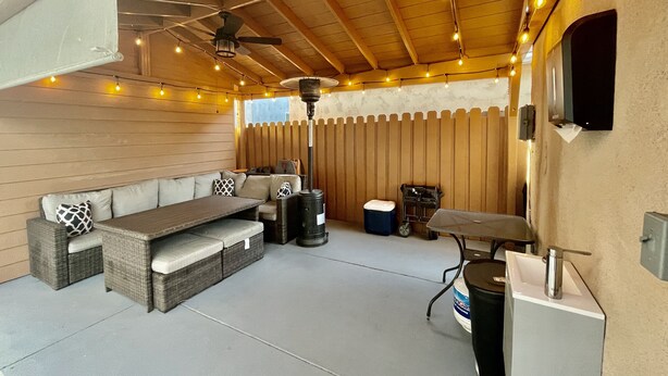 Ein möblierter Patio-Bereich im Freien mit Holzboden, Lichterketten, Deckenventilator, einer Sofagarnitur, einem Tisch, einem Grill und einem Mülleimer.