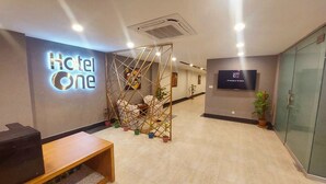 A hotel reception area with a 'Hotel One' sign, a reception desk, a seating area with a decorative partition, and a television screen displaying the hotel name and room count. - Hotel One Sargodha (Sargodha)