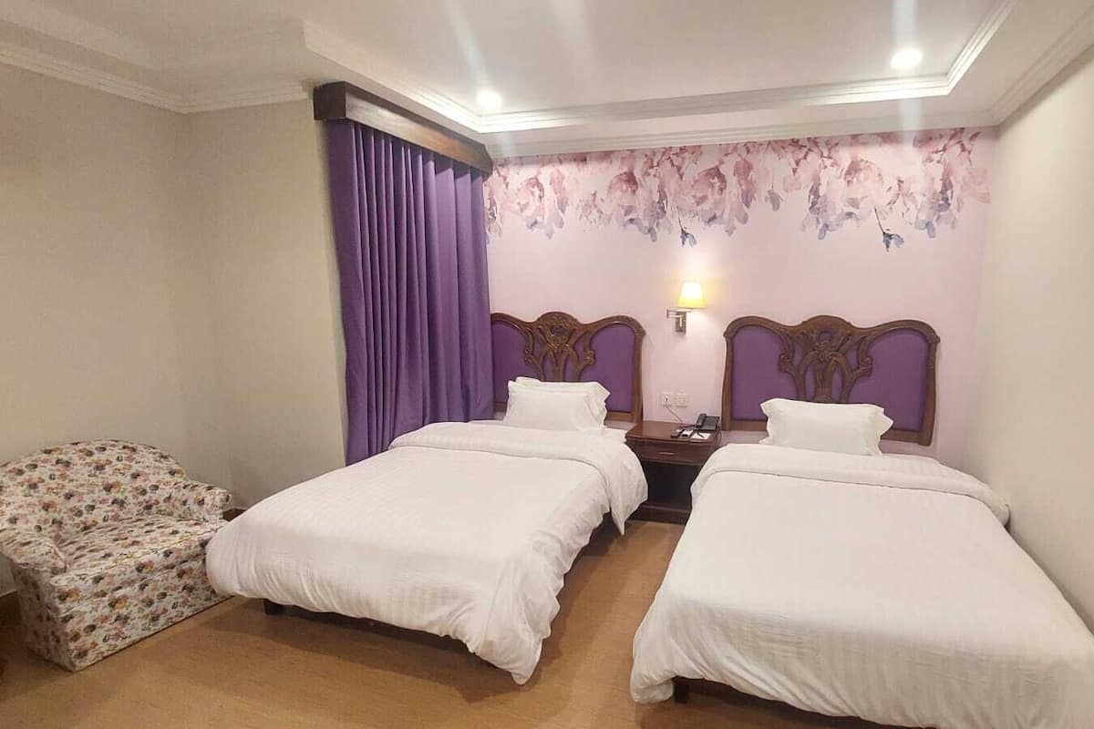 A hotel room with two beds, a nightstand, a lamp, and a floral-patterned chair.
