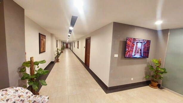 A hotel hallway with a long corridor, potted plants, framed pictures on the walls, and a television screen displaying a corridor scene. - Hotel One Sargodha (Sargodha)