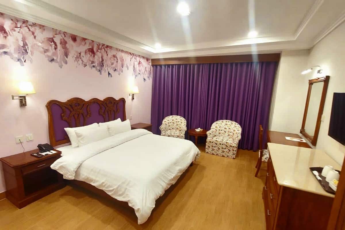 A hotel room with a large bed, two chairs, a desk with a mirror, and a purple curtain.