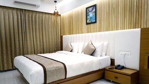 A modern bedroom with a large bed, wooden headboard, bedside tables, and a painting on the wall. - Hotel Tulsi (Surat)