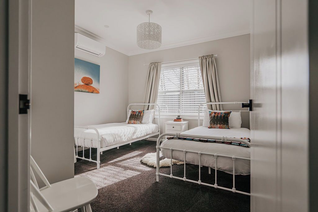 A bedroom with two single beds, a small bedside table, a chair, a window with curtains, and a hanging light fixture.