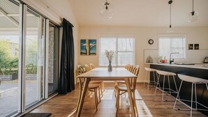 A modern dining area with a wooden table and chairs, a vase with flowers, and a kitchen area with bar stools.