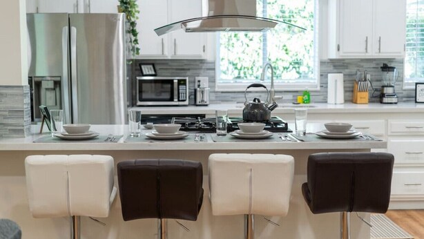 A modern kitchen with white cabinets, a central island with bar stools, a kettle, and a microwave.
