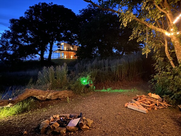 A modern house with a glass facade, surrounded by trees and a gravel path leading to a fire pit. - Tawny Lodge - 1 Bedroom - Blossom Farm (Haverfordwest)