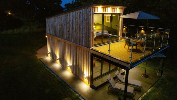 A wooden cabin with a glass balcony, outdoor seating, and a well-lit interior. - Tawny Lodge - 1 Bedroom - Blossom Farm (Haverfordwest)