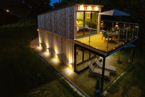 A wooden cabin with a glass balcony, outdoor seating, and a well-lit interior. - Tawny Lodge - 1 Bedroom - Blossom Farm (Haverfordwest)