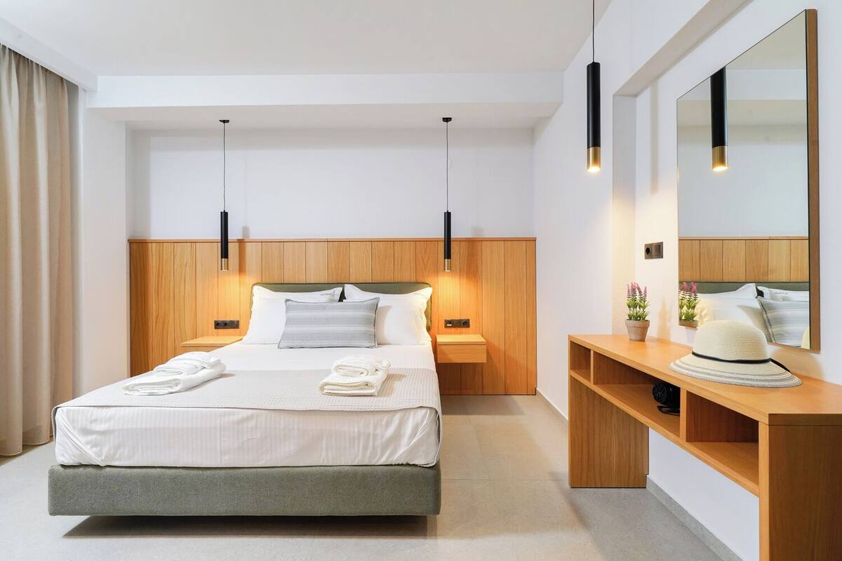 A modern hotel room with a large bed, bedside tables, a wooden headboard, a mirror, and a desk with a hat.