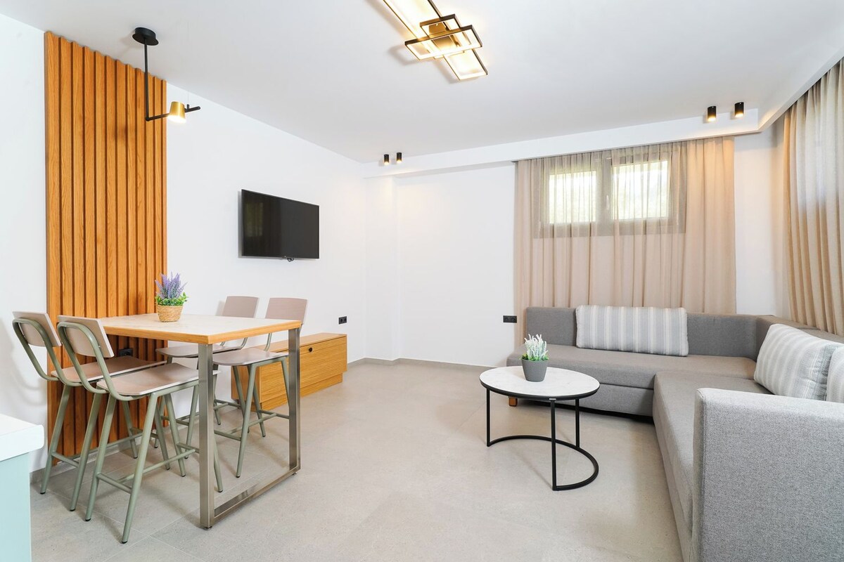 A modern living room with a dining area, a grey sofa, a small round table, and a TV mounted on the wall.