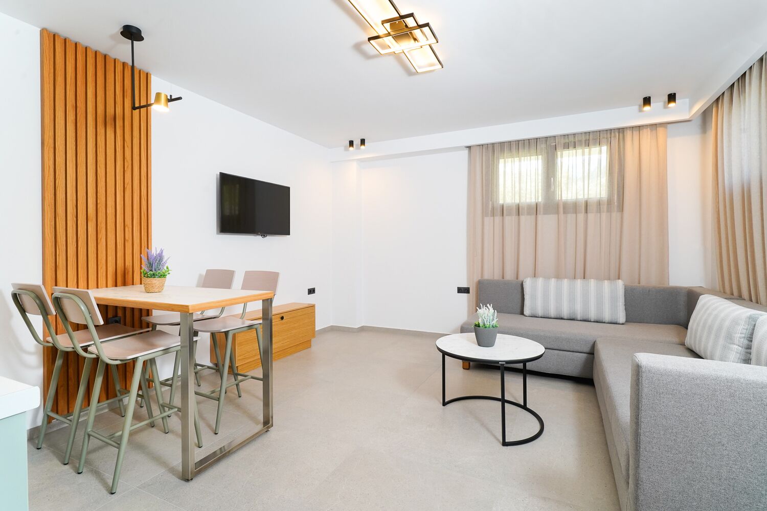 Ein modernes Wohnzimmer mit Essbereich, einem grauen Sofa, einem kleinen runden Tisch und einem an der Wand befestigten Fernseher.