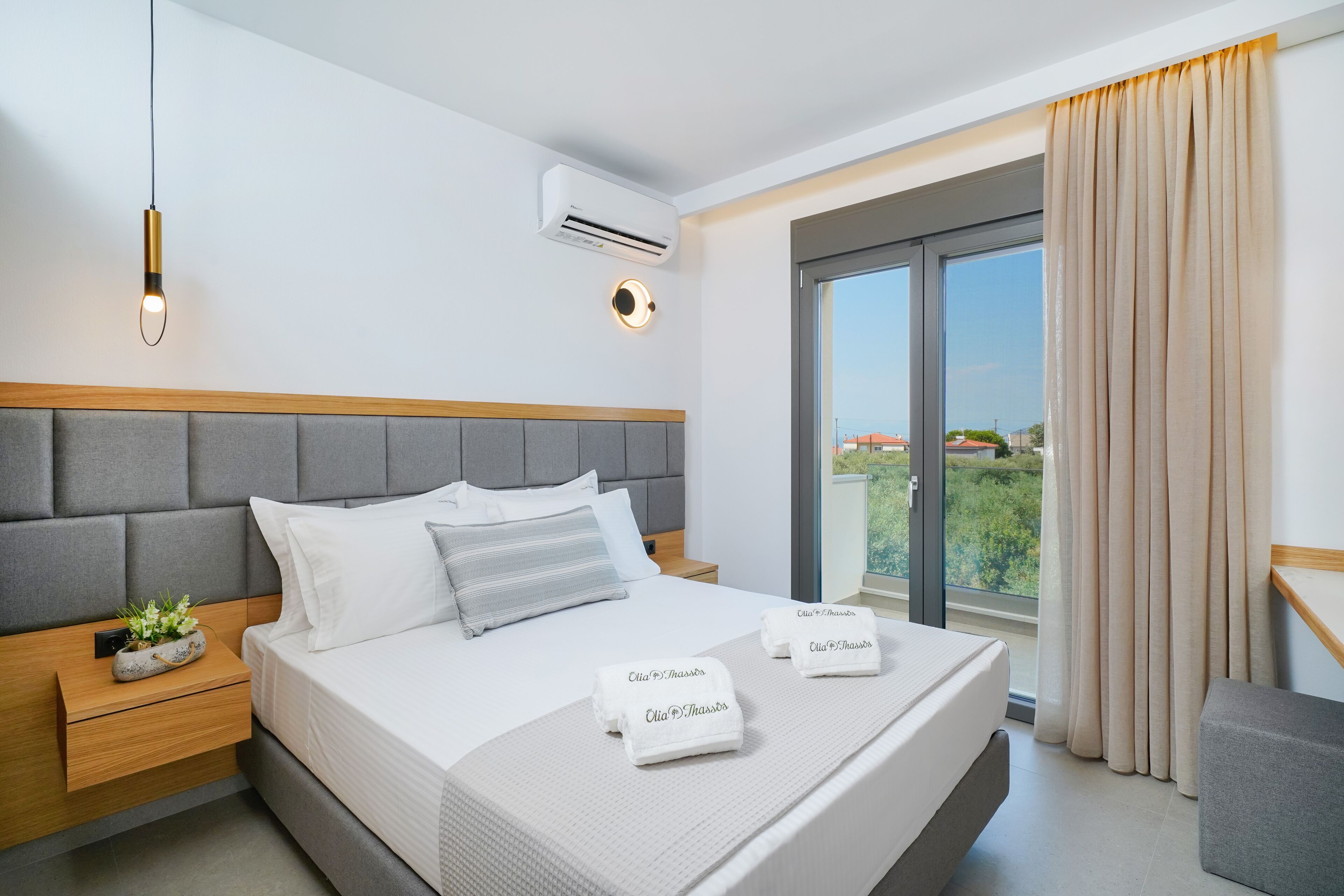 A modern hotel room with a large bed, grey headboard, bedside tables, a small plant, and a view of the outdoors through a large window.