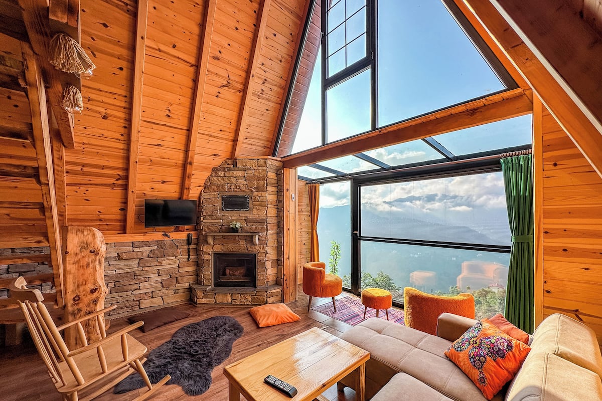 A cozy cabin interior with a stone fireplace, wooden furniture, and a large window offering a mountain view.