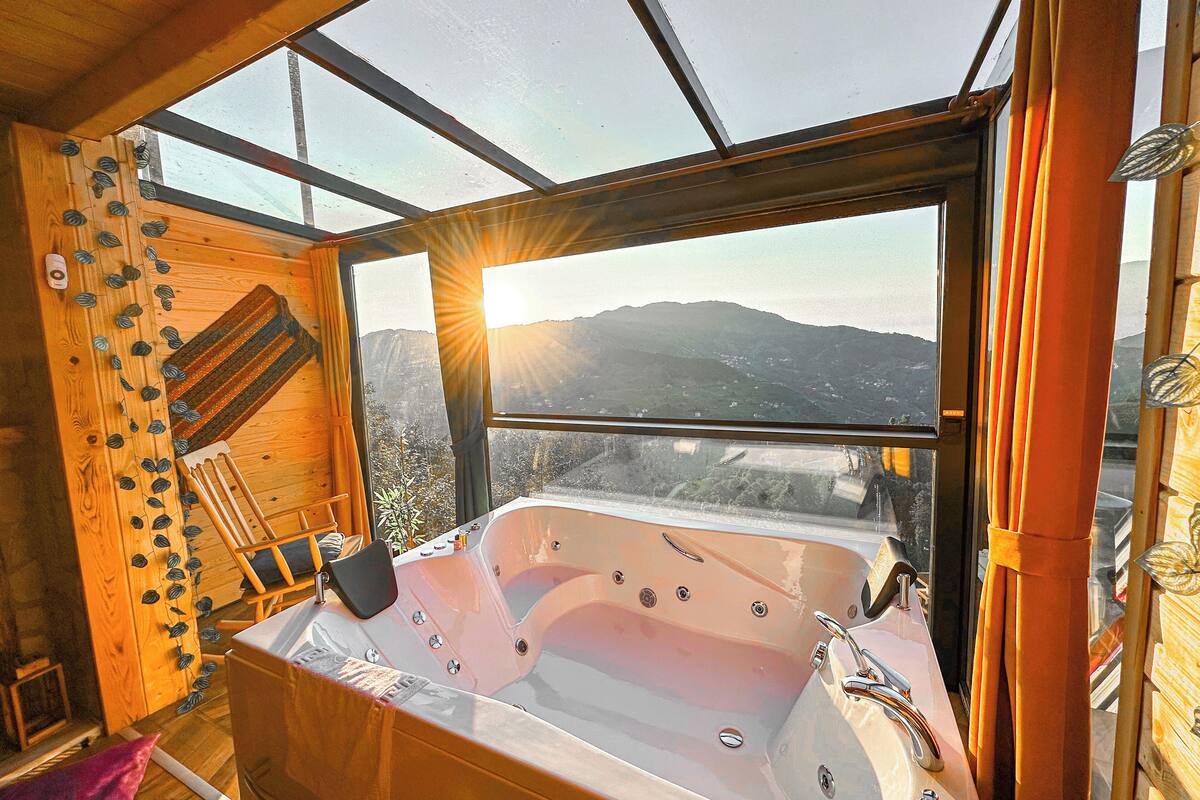 A wooden cabin with a large window, a bathtub, and a view of mountains.