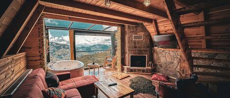 A cozy loft-style room with a stone fireplace, a large window offering a mountain view, a wooden ceiling, and a red leather sofa.