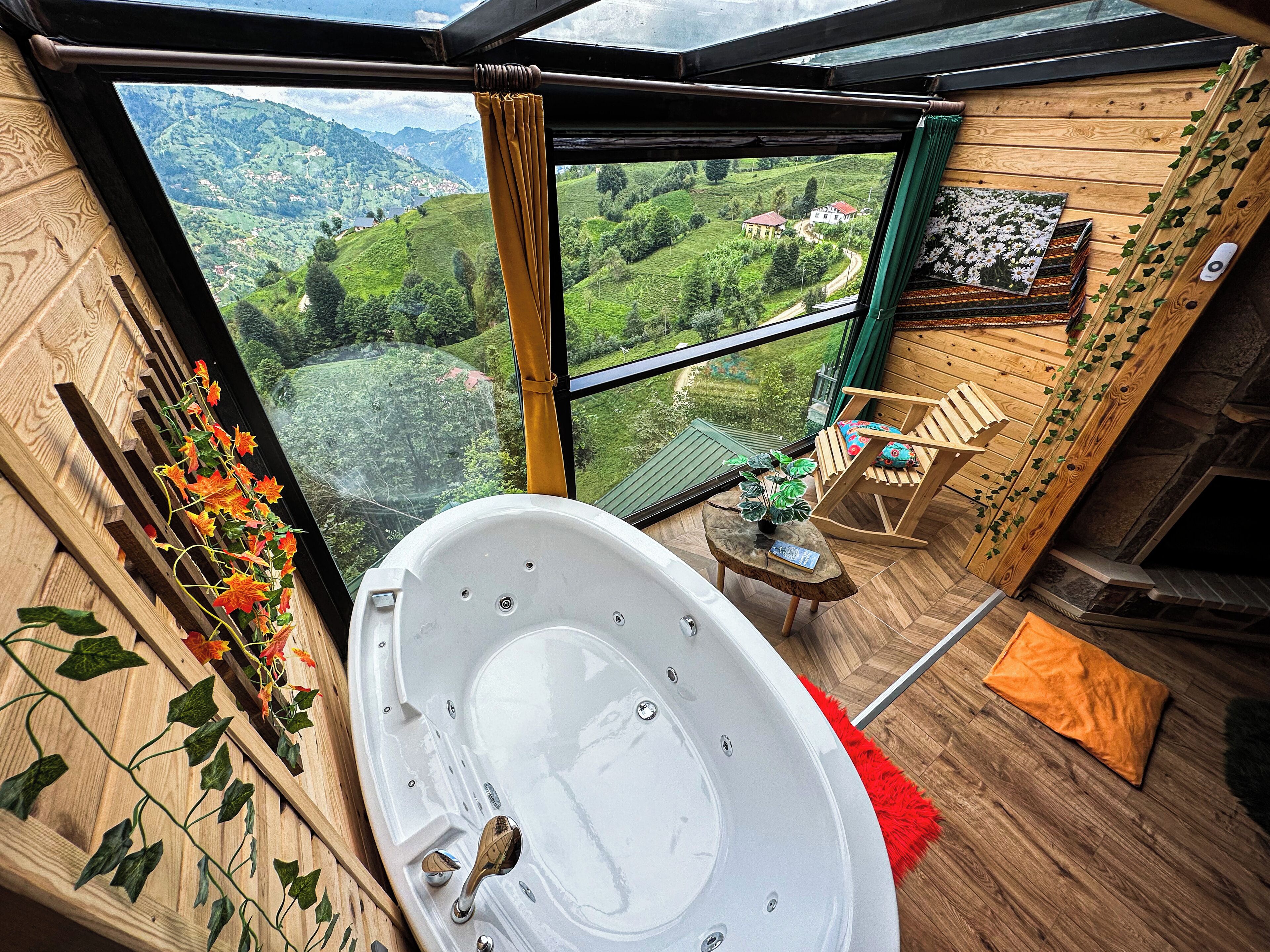 A room with a large bathtub, a wooden chair, a small table with a book, and a view of a green valley through a window.