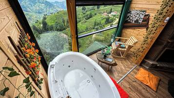 A room with a large bathtub, a wooden chair, a small table with a book, and a view of a green valley through a window.