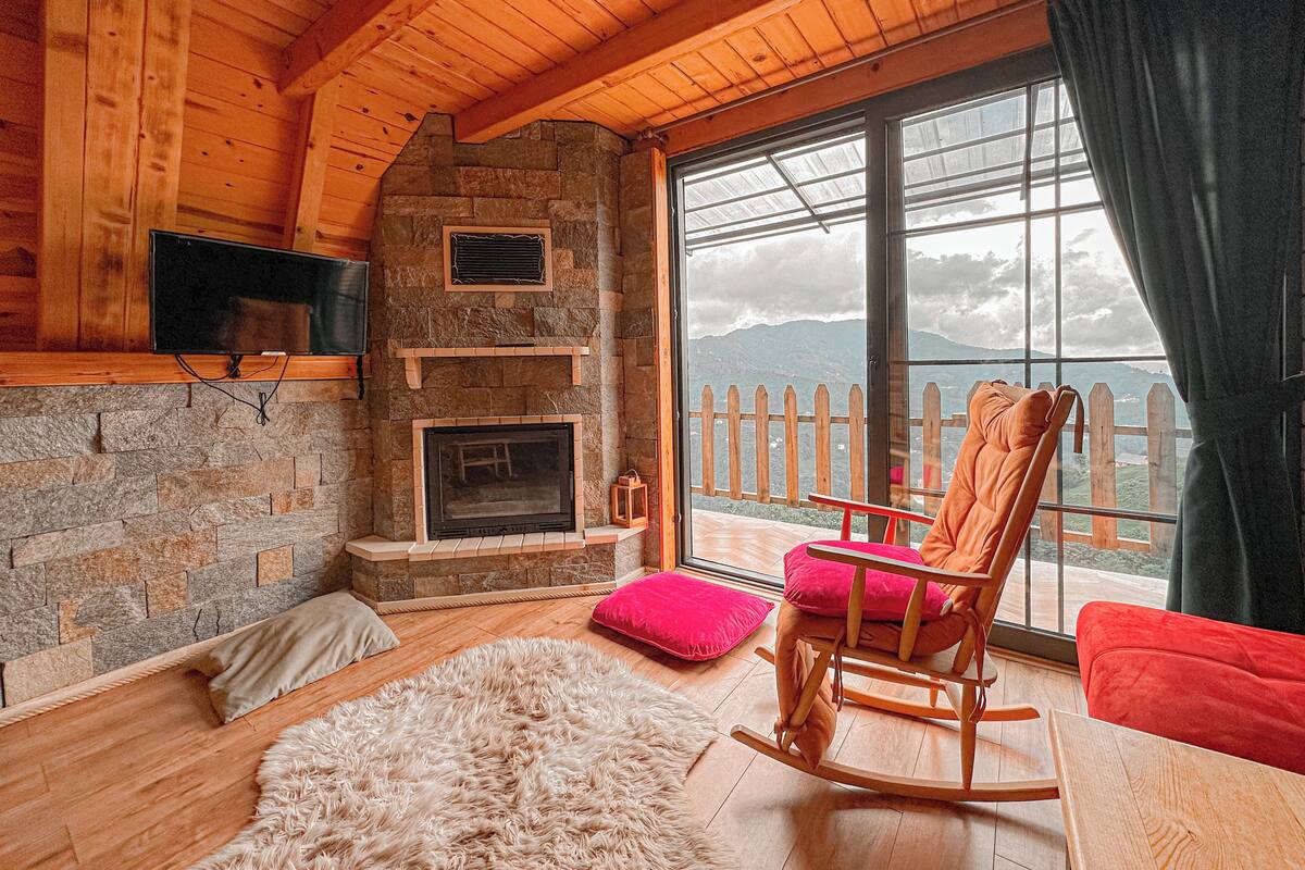 A cozy cabin interior with a stone fireplace, wooden walls, and a large window offering a mountain view.
