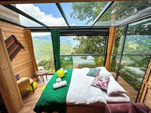 Une chambre avec un lit, une petite table et une vue sur un paysage verdoyant, visible à travers de grandes fenêtres.