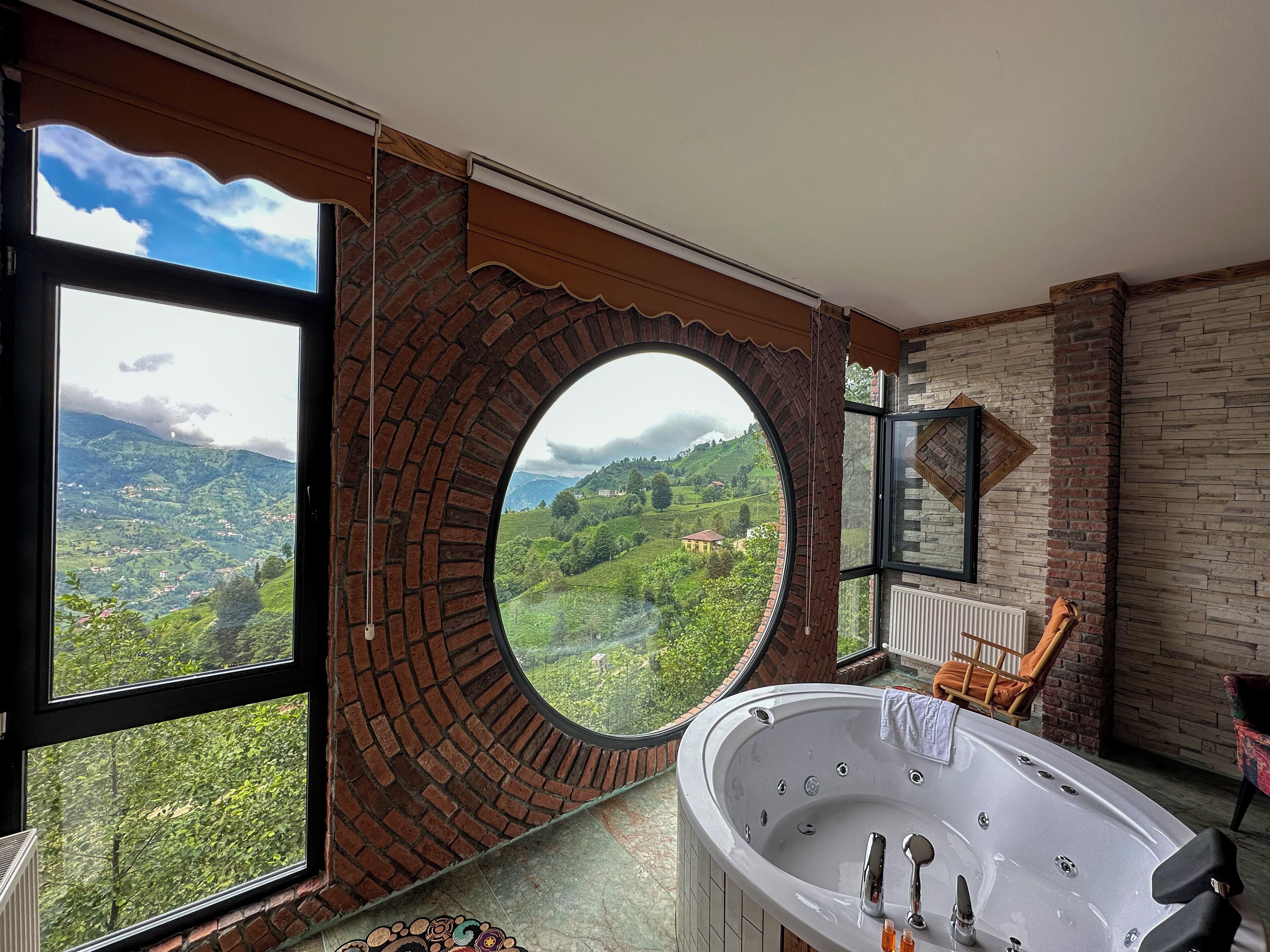 A bathroom with a large bathtub, a view of green hills through a circular window, and a brick wall.