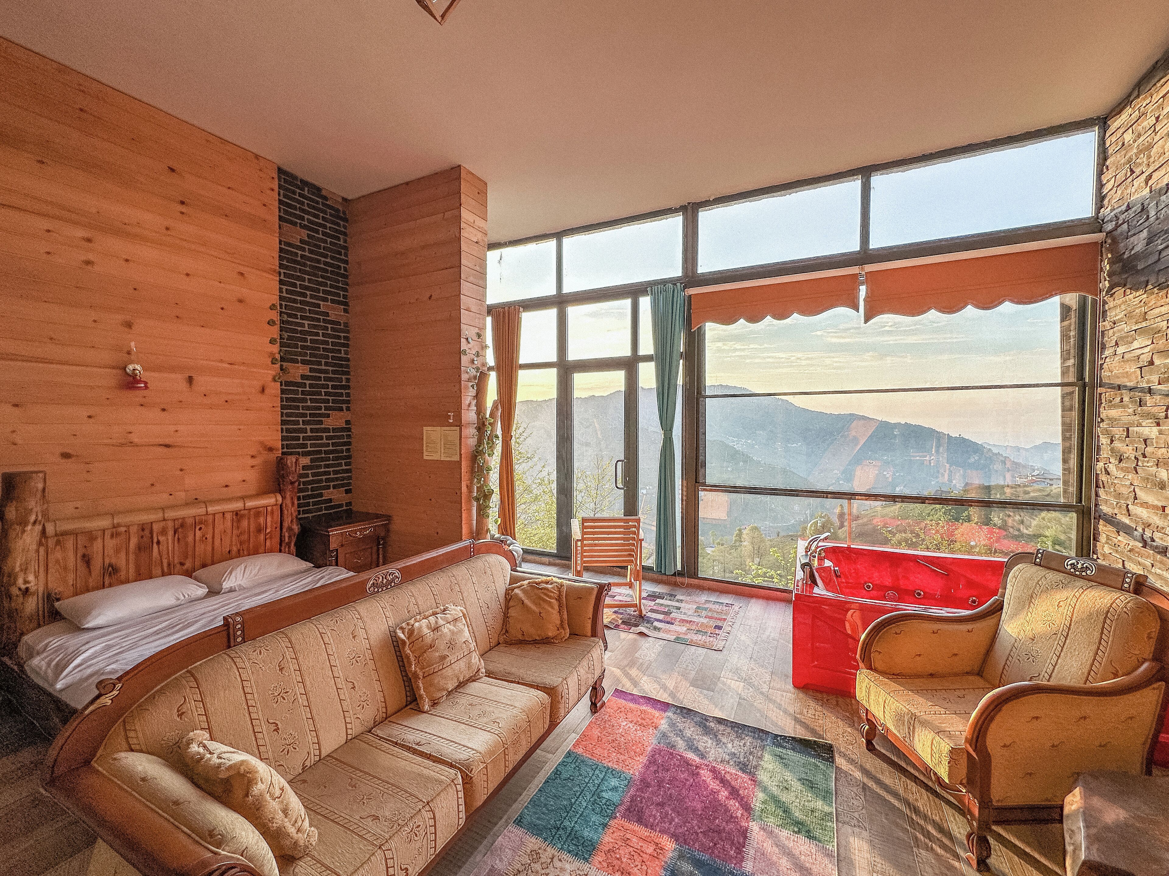 A cozy room with a wooden wall, a sofa, a red chair, and a view of mountains through large windows.