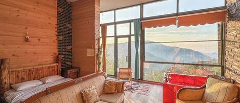 Quarto aconchegante com parede de madeira, um sofá, uma poltrona vermelha e vista para montanhas por grandes janelas.