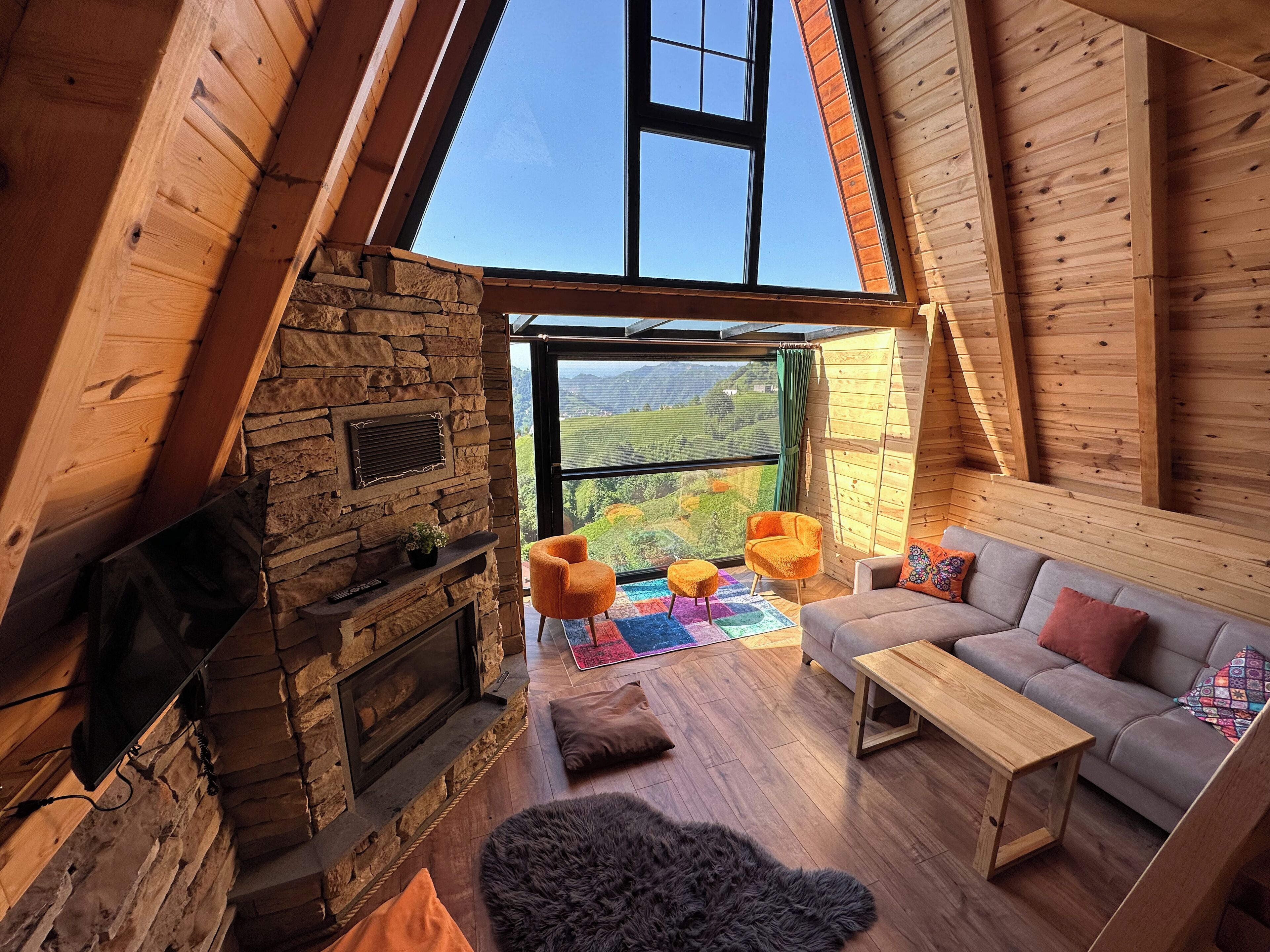 A cozy loft-style room with a stone fireplace, wooden beams, a sofa, and a large window offering a view of the countryside.