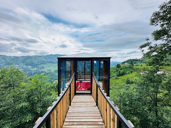Une terrasse en bois mène à une pièce fermée par des baies vitrées, où se trouve un fauteuil rouge, et d’où l’on a une vue sur un paysage montagneux.