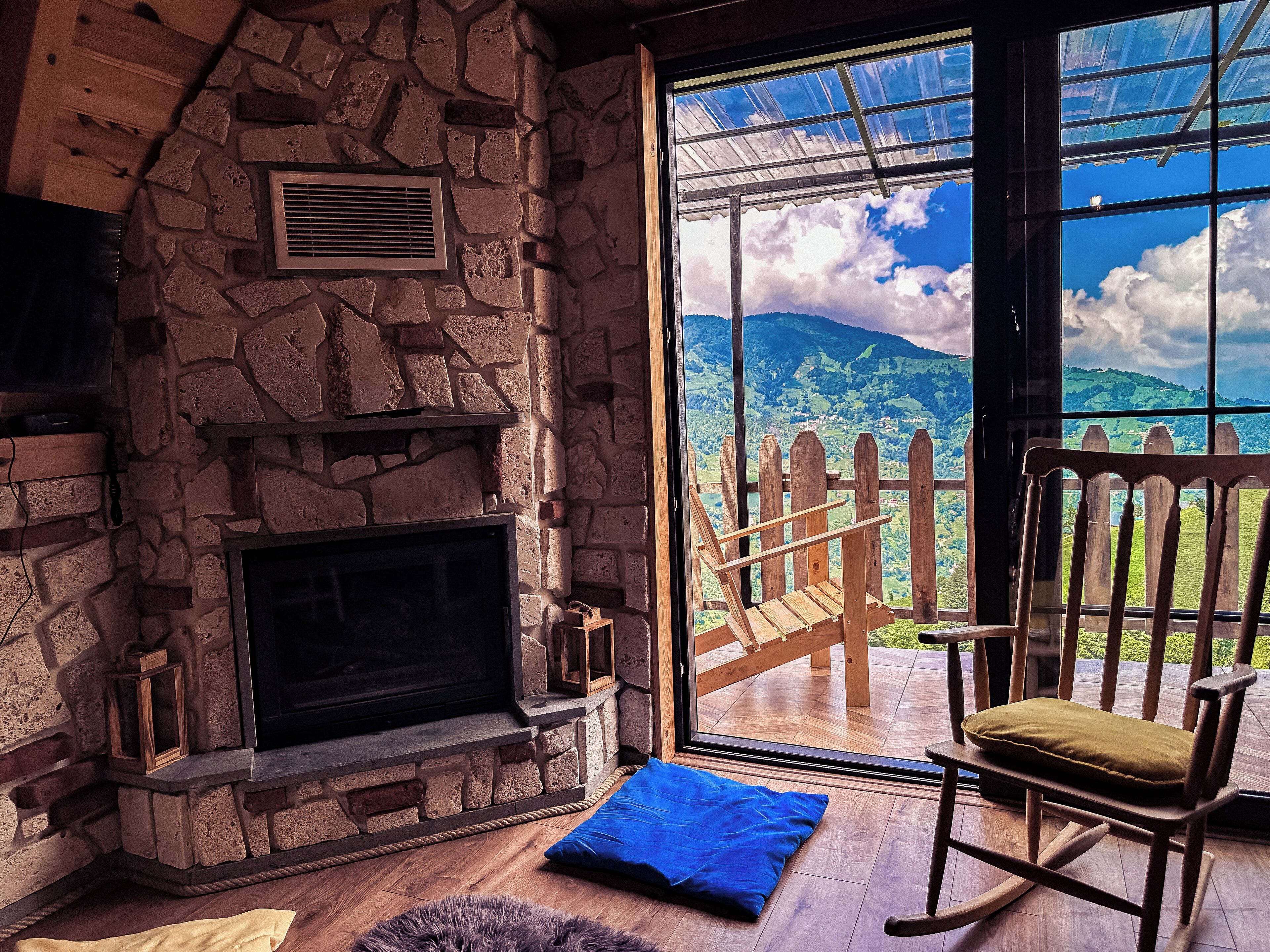 A stone fireplace, a wooden rocking chair, and a view of a mountainous landscape through a glass door.
