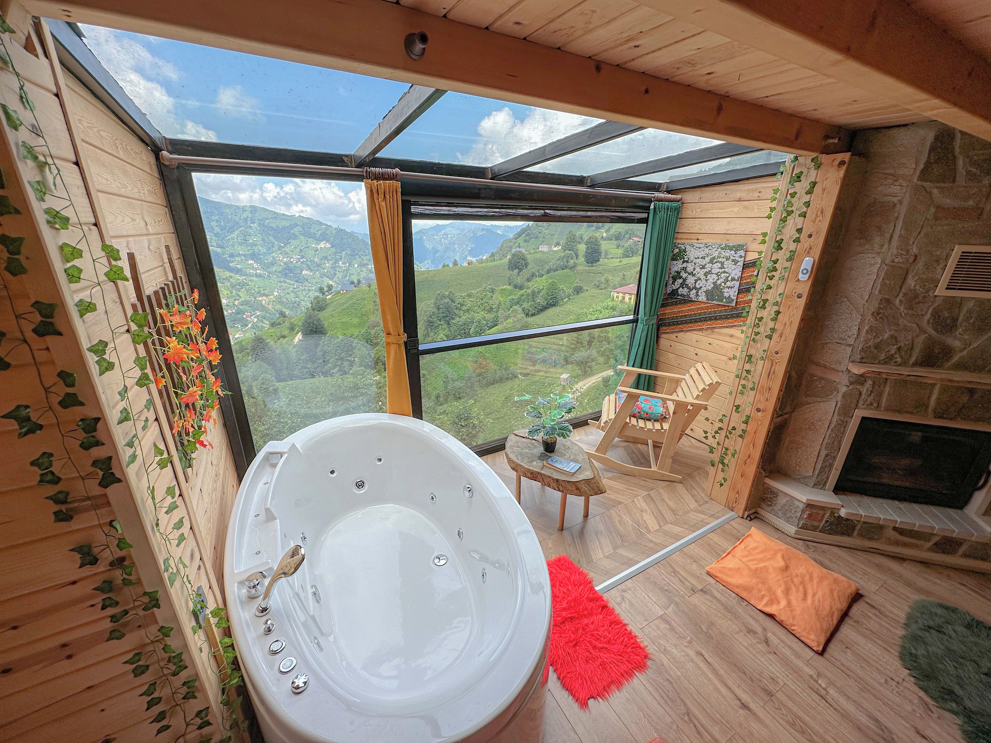 Un bagno con una vasca da bagno grande, una sedia di legno, un tavolino con una pianta e vista su un paesaggio montuoso attraverso una porta in vetro.