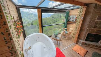 Un bagno con una vasca da bagno grande, una sedia di legno, un tavolino con una pianta e vista su un paesaggio montuoso attraverso una porta in vetro.