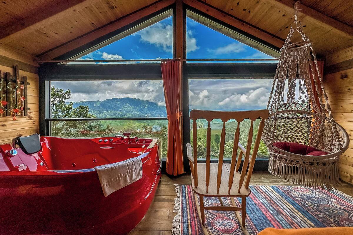 A room with a large window, a red boat, a wooden chair, and a hanging chair.