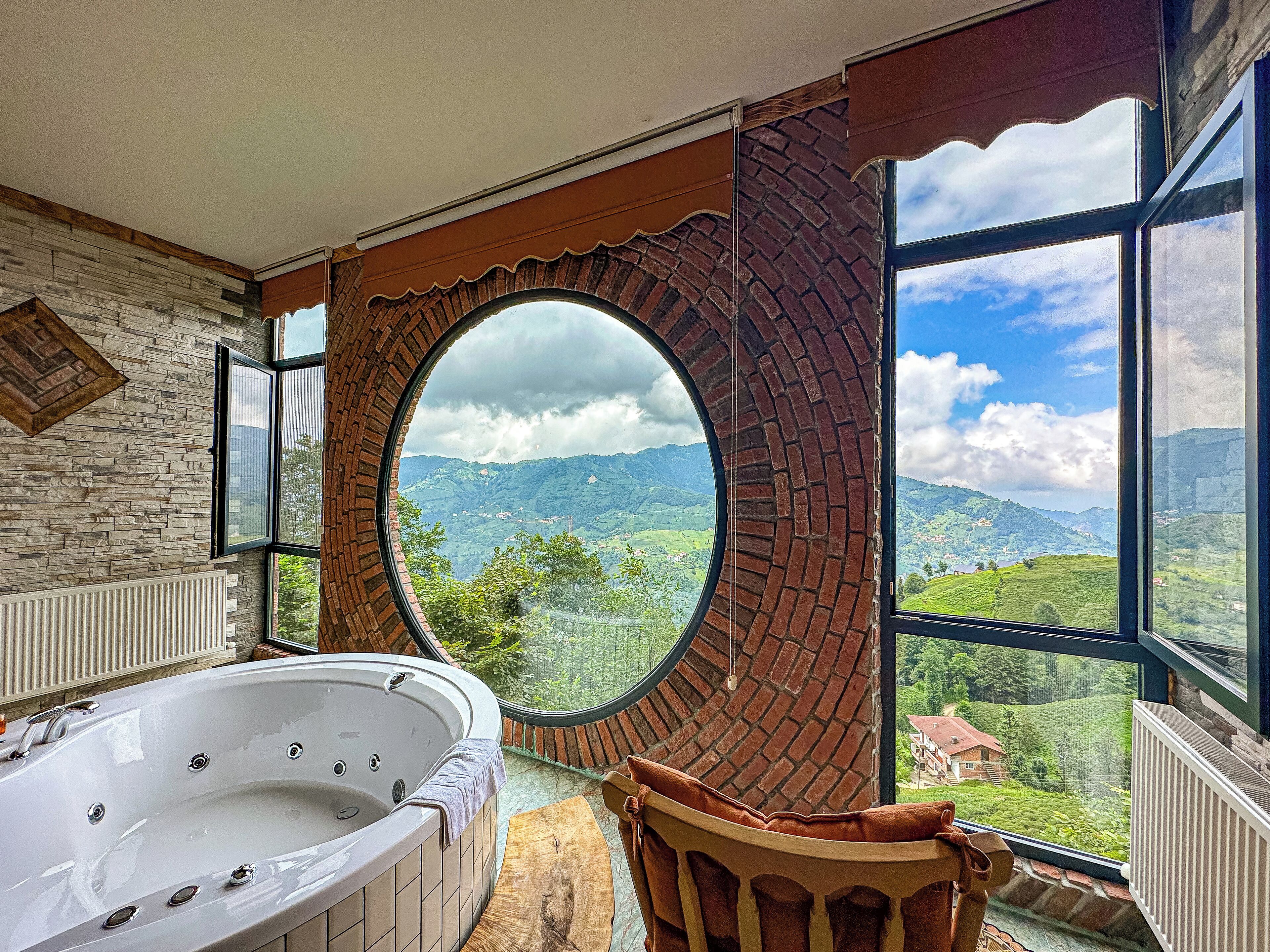 A bathroom with a large bathtub, a circular window with a brick arch, and a view of green hills.
