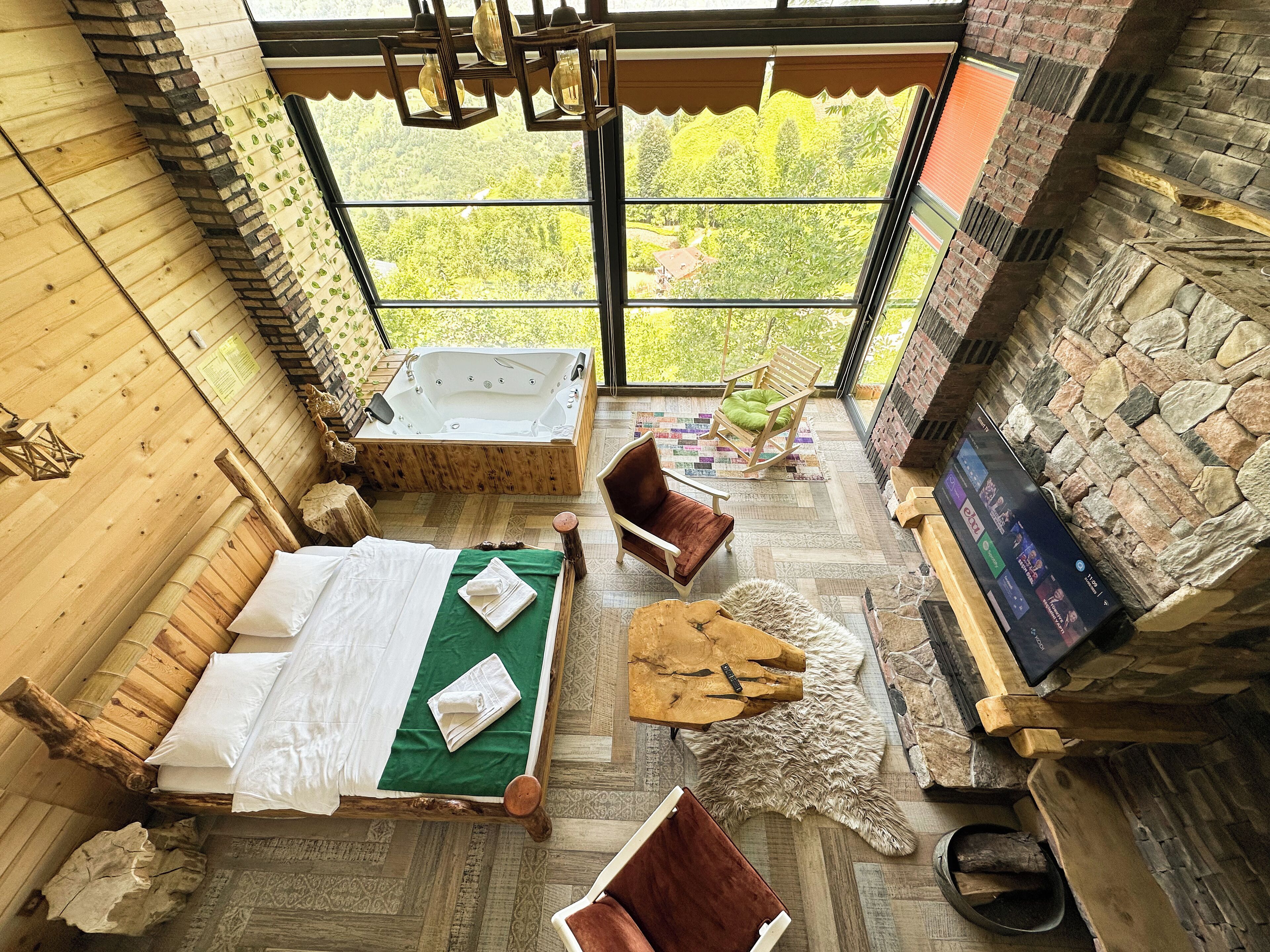 A cozy room with a wooden bed, a stone fireplace, a large window with a view of greenery, and a television on the wall.