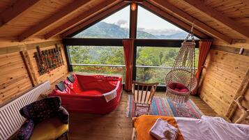 Une pièce chaleureuse avec un plafond en bois, un jacuzzi rouge, un fauteuil en osier, un lit recouvert d’une couverture blanche et orange, et une vue sur des collines verdoyantes.