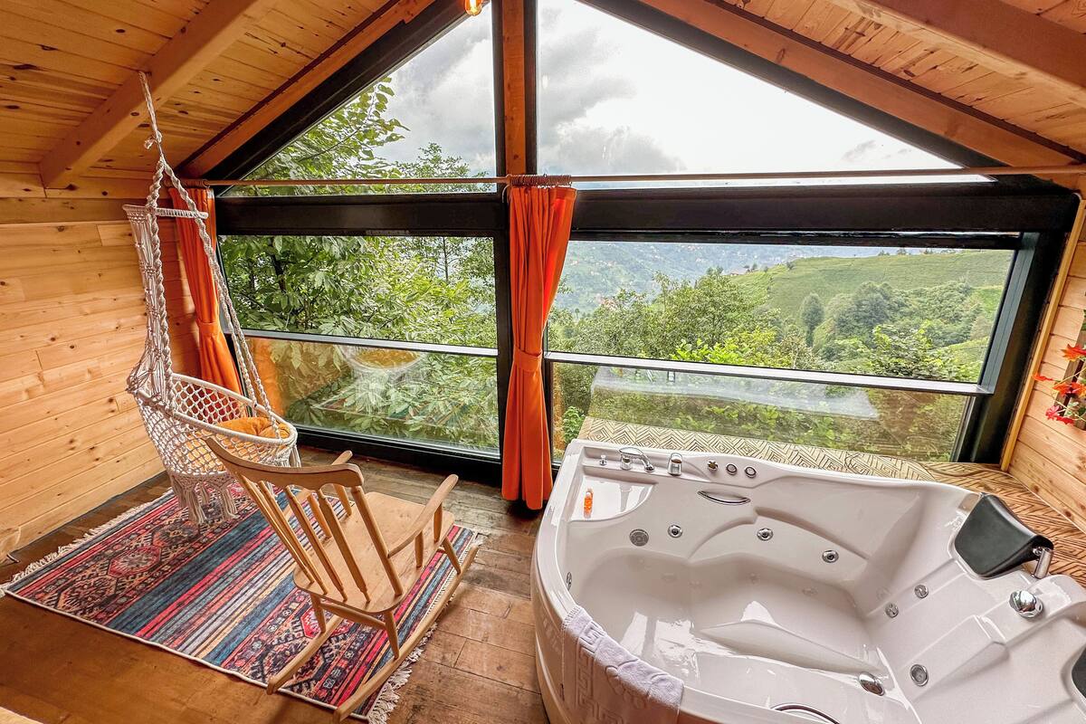 A wooden interior with a large window, a hanging chair, and a hot tub.
